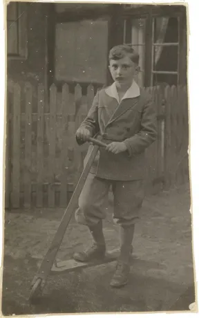 Walter Frankenstein&rsquo;s photograph as a child in fine clothing and standing in front of a fence with a scooter.
