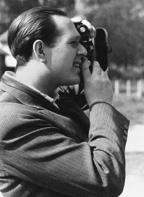 Die Aufnahme zeigt Fred Stein im Seitenprofil beim Fotografieren. Er tr&auml;gt ein Nadelstreifen-Jackett und ein Hemd ohne Krawatte.