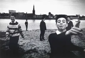 Schwarzwei&szlig;-Fotografie: Am Ufer eines breiten Flusses stehen sechs Jugendliche. Sie stehen nicht zusammen, sondern &uuml;ber die gesamte Bildfl&auml;che verteilt. Einige schauen zum Betrachter, andere hin&uuml;ber zur Stadt, die sich auf der gegen&uuml;berliegenden Seite des Flusses entlang zieht. Die Gruppe macht einen ausgelassenen Eindruck.
