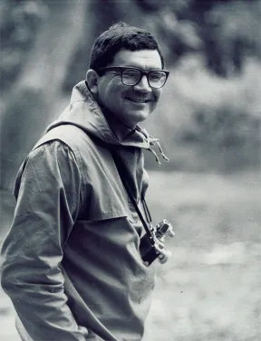 Schwarzwei&szlig;-Fotografie eines Mannes (Leonard Freed), der in die Kamera blickt und l&auml;chelt. Zu sehen ist sein Oberk&ouml;rper, er tr&auml;gt Jacke und Brille sowie eine Kamera um den Hals. Er steht im Freien, die Landschaft im Hintergrund ist unscharf.