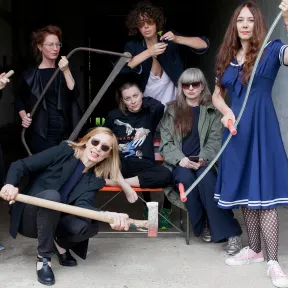 Group photo of the musicians from &ldquo;Monika Werkstatt&rdquo;. Some of the woman present each one of the following objects: an axe, a sickle, a saw and a flower.