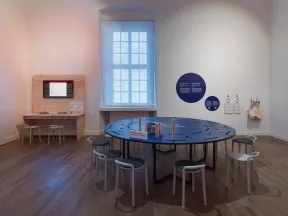 A view of an exhibition space featuring a large round table surrounded by stools, with recesses in the table for inserting wooden pieces. In the background is another interactive station with a screen and tablets.
