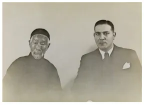 Schwarz-Wei&szlig; Bild. Links ein &auml;lterer Mann in dunkler Kleidung. Rechts ein mittelalter Mann in einem Anzug. Beide schauen in die Kamera. 