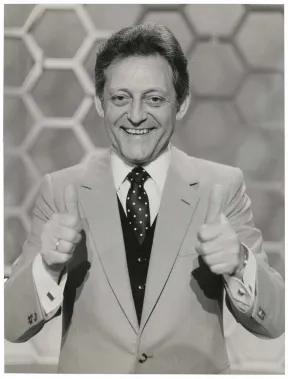 The black and white photo shows a half-portrait of a man in suit and tie with both thumbs up and looking friendly into the camera, with honeycomb pattern in the background. 