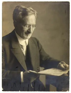 Schwarz-Wei&szlig;-Fotografie eines Mannes im Anzug und mit Brille, zeitungslesend an einem Tisch sitzend.