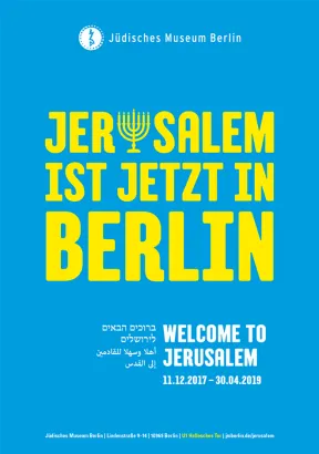 Ausstellungsplakat mit Aufschrift "Jerusalem ist jetzt in Berlin" in gelben Buchstaben auf blauem Hintergrund. Der Buchstabe 'u' in Jerusalem hat die Form einer Menora