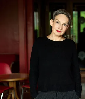 Portr&auml;tbild von Marion Brasch mit H&auml;nden in den Hosentaschen und Bartisch im Hintergrund