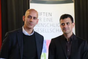 Portr&auml;tfoto von zwei M&auml;nnern in Frontalansicht. Sie l&auml;cheln und tragen Anz&uuml;ge, aber keine Krawatten.