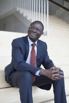 Karamba Diaby is sitting on a staircase, wearing a blue suit with a red check tie.