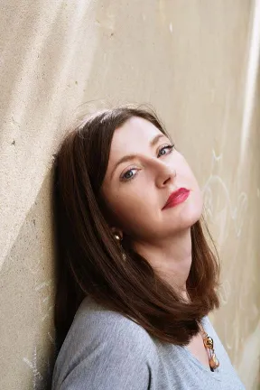 Woman with long brown hair and red lipstick leans her head against a wall and looks at the camera