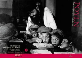 The black and white photo shows several boys in caps sitting close together at a table. In front of them are open books, behind them stands a man and points to a line of text