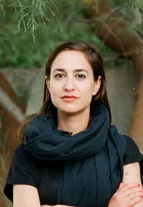 A woman with long brown hair stands in front of a green background and looks into the camera.