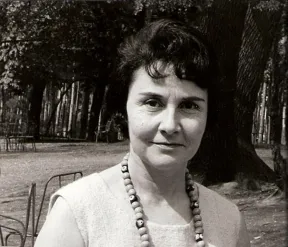 Schwarz-Wei&szlig;-Foto einer mittelalten Frau mit kurzen dunklen Haaren und auff&auml;lliger Kette um den Hals, die freundlich-skeptisch in die Kamera schaut, im Hintergrund B&auml;ume.
