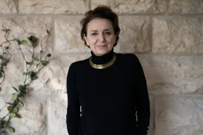 A short-haired woman wearing a black turtleneck sweater with a gold necklace stands in front of a light-colored stone wall with ivy vines and looks into the camera.