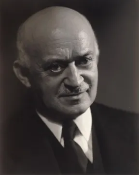 Black and white photograph of a man with a moustache and bald head in a suit, shirt and tie.