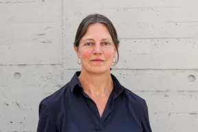 Eine Frau mit zusammengebundenen Haaren und in dunkelblauer Bluse steht vor einer hellen Betonwand und blickt geradeaus in die Kamera.