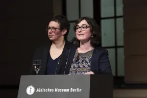 Alina Gromova steht an einem Pult auf dem das Logo und der Schriftzug des J&uuml;dischen Museums stehen. Auf dem Pult steht ein Wasserglas. Hinter ihr steht Susan Kamel.