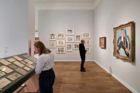 Ausstellungsraum mit Besucher*innen und Gem&auml;lden an wei&szlig;er Wand, einer Vitrine mit Gem&auml;lden und Zeichnungen in der Mitte des Raumes.