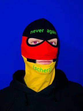 Portait Foto einer maskierten Person vor blauem Hintergrund. Die Person tr&auml;gt eine schwarz-rot-gelbe Sturmhaube mit der Aufschrift never again auf der Stirn sowie (scherz) auf dem Kinn.