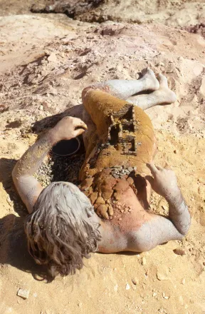 Filmstill aus dem Film &bdquo;Birth&ldquo; von Charles Simmonds: Eine unbekleidete, mit Sand und Erde bedeckte Frau liegt auf einem sandigen Boden. 