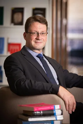 Ein Mann mit dunklem Haar, Brille, Anzug und Krawatte schaut freundlich in die Kamera.