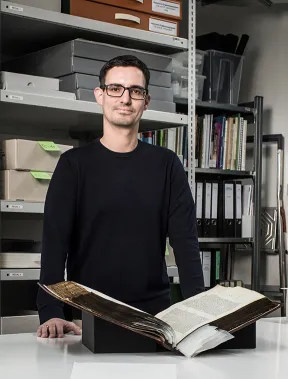 Ein Mann steht hinter einem Arbeitstisch in einem Atelier; vor ihm liegt ein altes aufgeschlagenes Buch