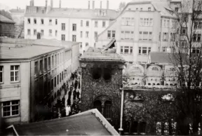 Das Fotot zeigt eine zerst&ouml;rte Synagoge, deren Dach fehlt, es scheint verbrannt zu sein, Tr&uuml;mmer liegen auf dem noch bestehenden Eingangsportal, in der Sta&szlig;e davor befinden sich viele Passanten.  