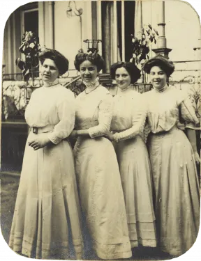 Black and white photo showing four young ladies standing in a row diagonally behind each other in three-quarter profile in front of a house. All are wearing white, high-necked dresses and updos.