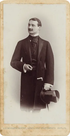 Black and white portrait photo of a standing young man. The body is seen frontally, the head turned to the left in three-quarter profile. He wears a mustache and a frock coat, in his hand he holds a top hat and a walking stick.