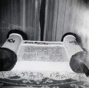 Schwarz-wei&szlig;-Foto eines Kuchens in Form einer Tora-Rolle auf einem Tisch mit Spitzentischdecke