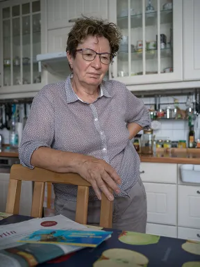 Eine Frau mit Brille und gepunkteter Bluse steht auf eine Stuhllehne gest&uuml;tzt in einer K&uuml;che