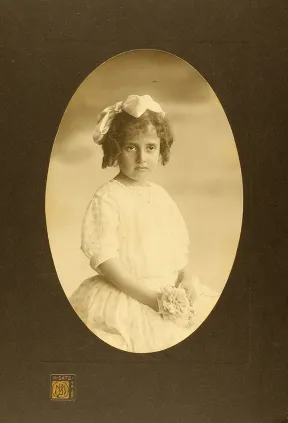Studioaufnahme eines M&auml;dchens in wei&szlig;em Kleid, mit Schleife im Haar und Stoffblume in der Hand