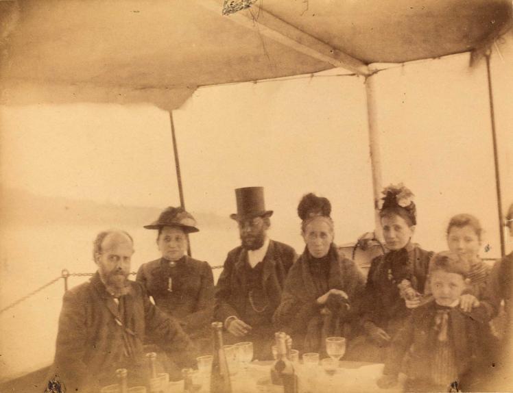 Yellowed photo of festively dressed people at a table with wine glasses and bottles on a boat trip, lake in the background.