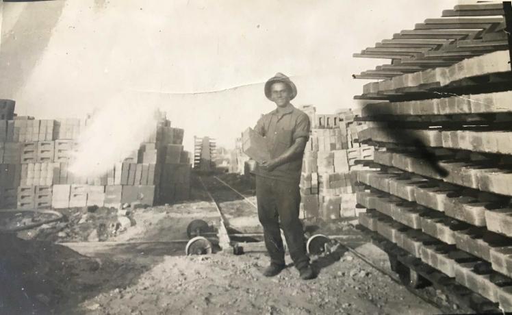 Black-and-white photo of a young man in a hat with bricks in his arms; more bricks in the background