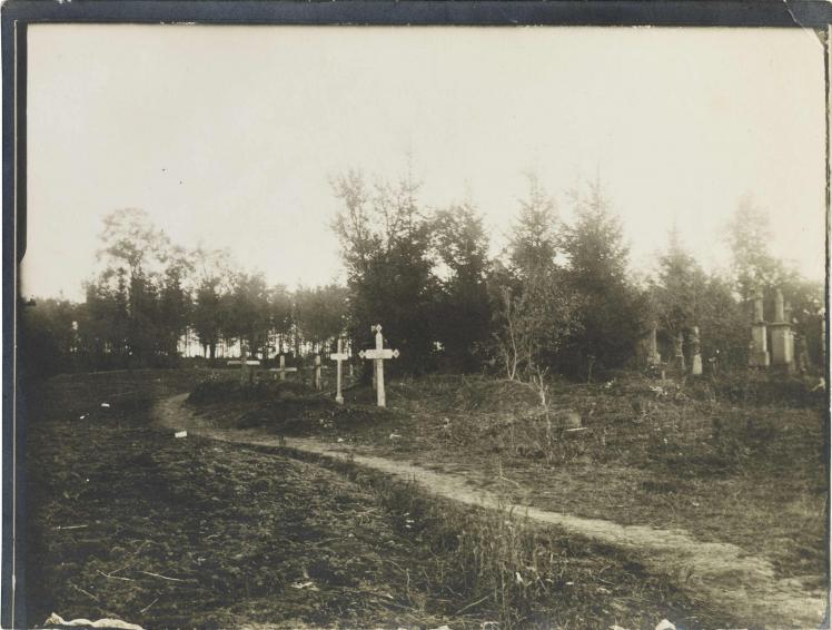Black-and-white photograph: Graves with crosses near a sandy path, with more graves and a forest in the background