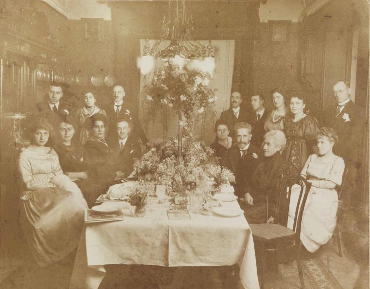 Schwarz-weiß-Foto einer Gruppe mit festlich gekleideten Personen, die in einer bürgerlichen Wohnung um einen gedeckten Tisch versammelt sind