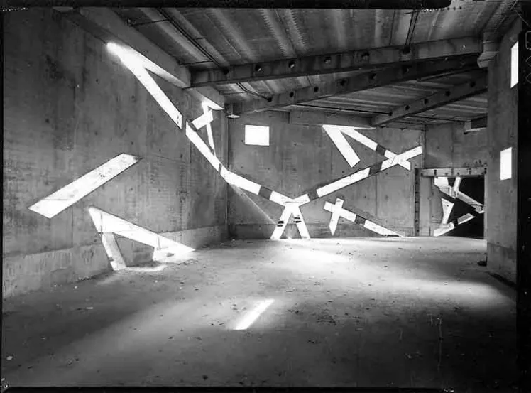 Die Schwarz-Wei&szlig;-Fotografie zeigt die Innenraumaufnahme eines Geb&auml;udes im Aufbau. In den Betonw&auml;nden sind schr&auml;g verlaufende Fenster, durch die Licht einf&auml;llt.