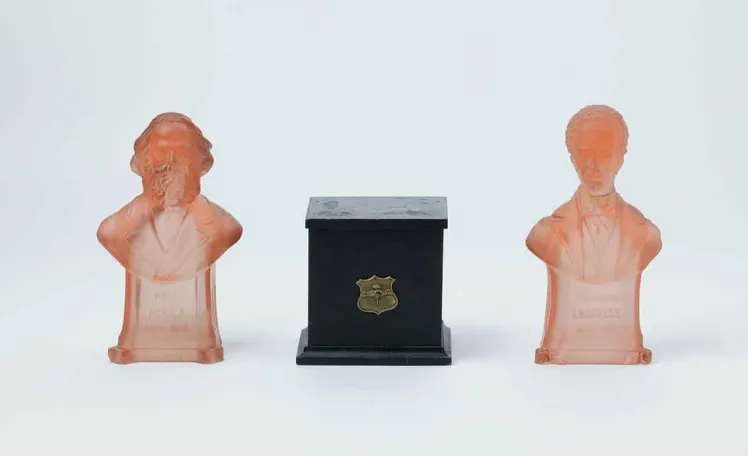 Two busts made of red, transparent glass. The one on the left depicts Karl Marx, the one on the right Ferdinand Lassalle. Between them is a dark brown wooden music box.