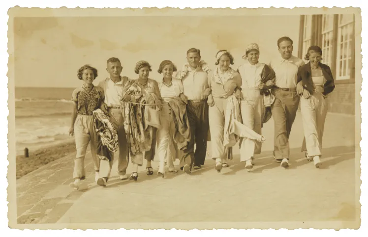 Old black-and-white photo. A group of cheerful young adults, men and women, walking in a row arm in arm along the beach.