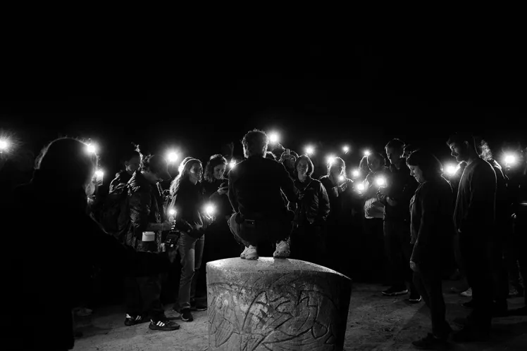 Eine Gruppe von Menschen mit Taschenlampen stehen im Dunkeln und schauen auf eine Person, die vor ihnen auf einem Stein hockt und der Kamera den Rücken zudreht.
