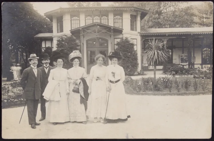 Schwarz-Weiß Bild. Zwei Männer in dunklen Anzügen stehen links. Rechts daneben stehen vier Frauen in hellen Kleidern. 