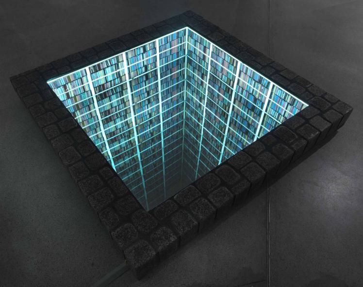 The view from above into a library sunk into the ground with a square plot; the shelved walls are illuminated green-blue.