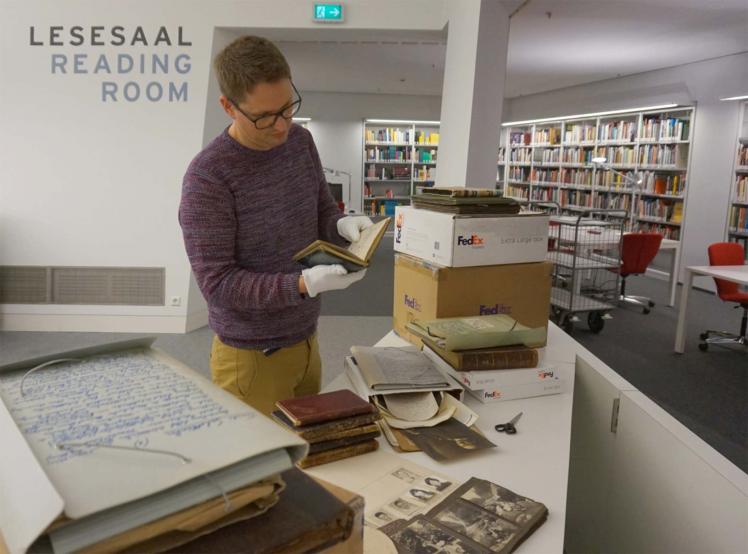 Partially unpacked box in a library. The table in the foreground is covered in black-and-white photos and other documents; an Archive employee is reading one of the unpacked albums