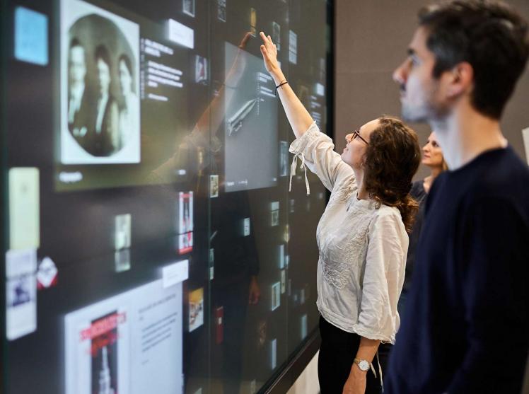 A woman and a man stand in front of a wall of large screens on which numerous are depicted. The woman points to one of the objects.