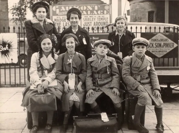 Black-and-white group photo of seven children in coats with baggage.