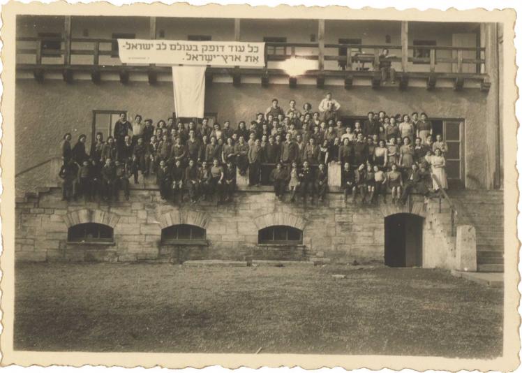 Auf der Schwarz-Weiß-Fotografie sitzen circa 100 Frauen und Männer auf einer Veranda. Über ihnen hängt ein Banner mit einem hebräischen Zitat aus einem Lied von Pooa Grinshpon.