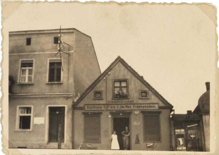 Die Aufnahme zeigt ein Wohnhaus. Über der Eingangstür ist ein Schild mit der Aufschrift „Schankwirtschaft // Kaufhaus S. Klein jr. Inh. Max Frankenstein“. In der Eingangstür stehen eine Frau im hellen Kleid (Martha Frankenstein) und ein junger Mann.