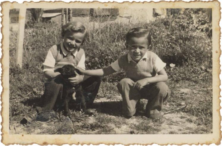 Auf der Schwarz-Weiß-Fotografie hocken die beiden Kinder in einem Garten und streicheln einen Hundewelpen.