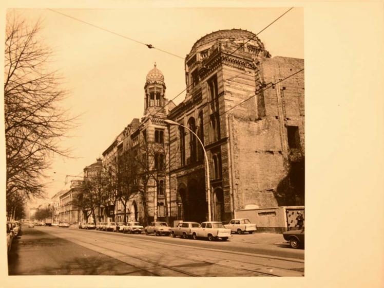 Die Fotografie zeigt die Neue Synagoge in der Berliner Oranienburger Straße im zerstörten Zustand während der DDR. Die Oranienburger Straße ist mit Autos des Typ Trabant flankiert.