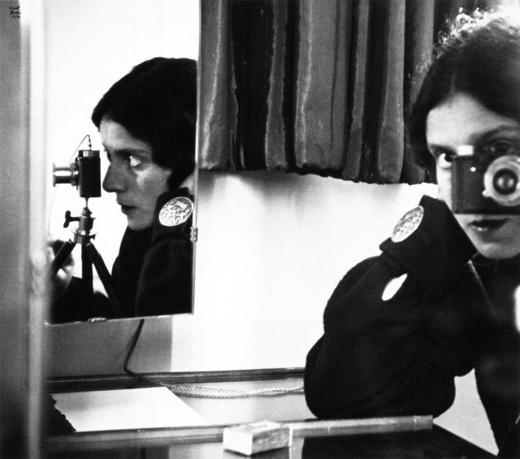 Schwarz-Weiß-Fotografie einer Frau, die durch eine Kamera sieht, ein Spiegel zeigt sie zusätzlich im Profil